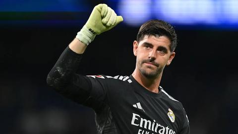 Same goalkeeper in black Real Madrid kit raising a gloved fist, dark stadium background; only kit sponsor on shirt