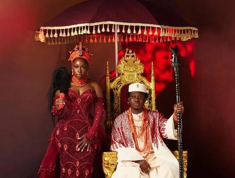 Bride in red beaded gown and groom in white agbada seated on a gold throne under a fringed umbrella