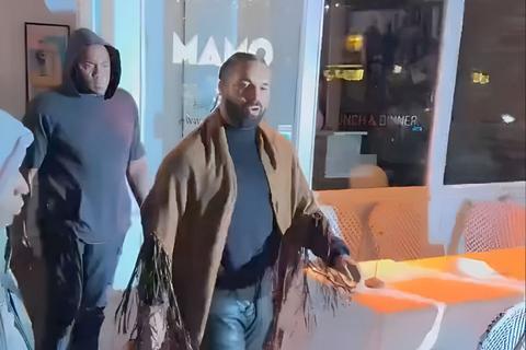 bearded man in a fringed shawl walking past a restaurant window at night, another man behind him