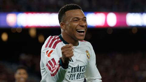 Real Madrid forward in white kit celebrating, fists clenched, smiling; sponsor wording on shirt is part of the kit