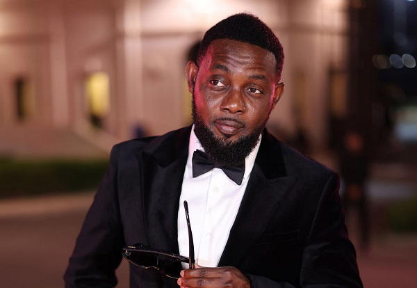 AY Makun in a black tuxedo and bow tie, holding sunglasses, outdoors at night