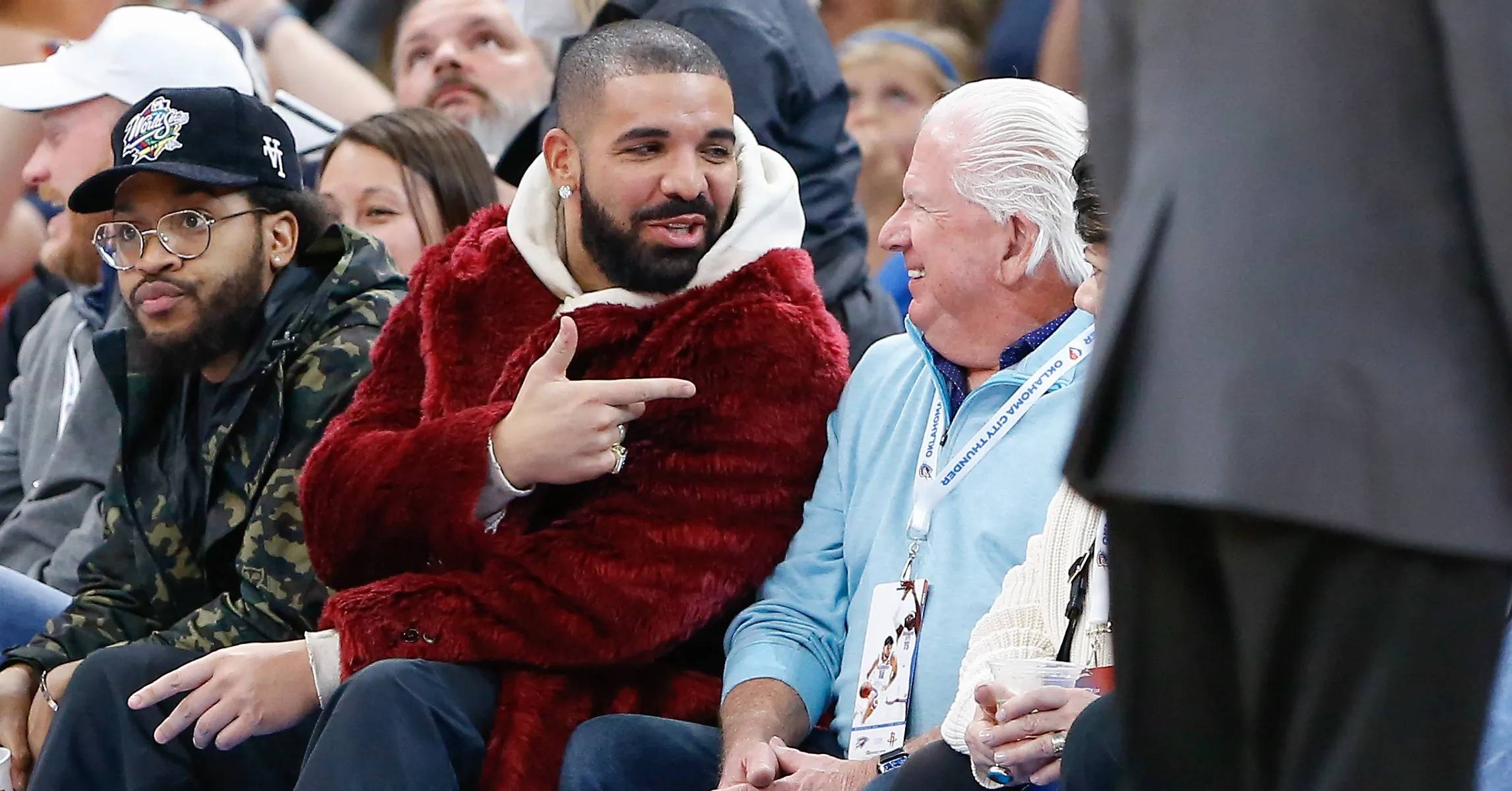 Drake in a red fur coat sitting courtside at a basketball game, pointing; not the lawsuit moment
