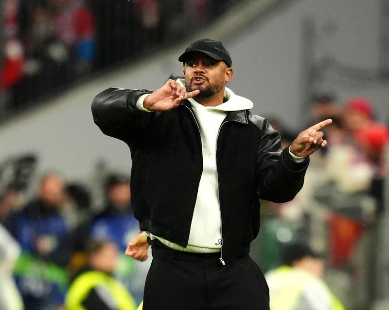 Man in black cap and leather jacket over white hoodie gesturing on the touchline, blurred stand behind