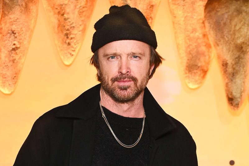 Bearded man in a black beanie and coat posed against a warm orange backdrop