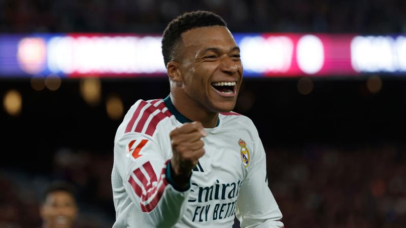 Real Madrid forward in white kit celebrating, fists clenched, smiling; sponsor wording on shirt is part of the kit