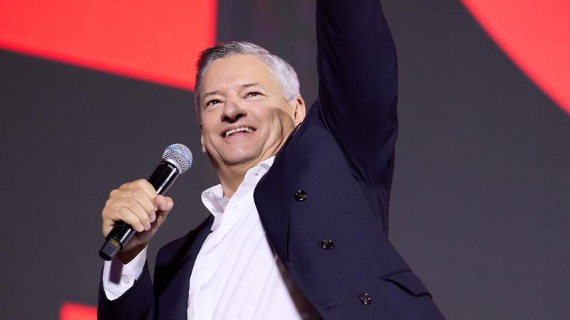 executive with grey hair speaking into a handheld mic, one arm raised, on a red-lit stage