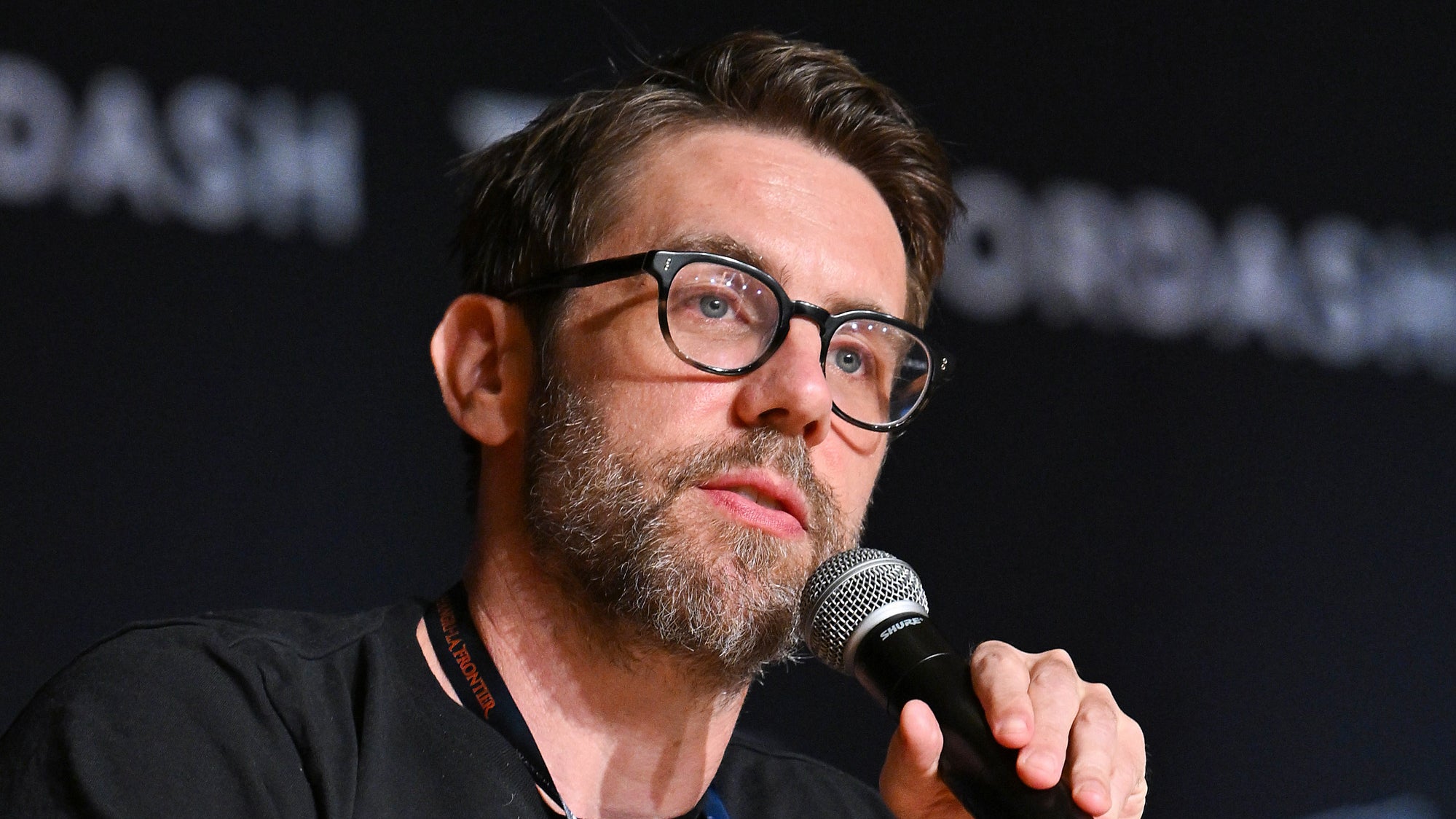 David Bruckner speaking into a microphone onstage at a horror panel, dark backdrop with large white lettering
