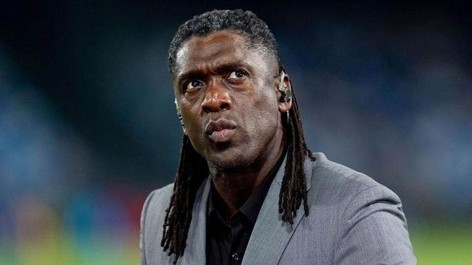 Close-up portrait of a man with long dreadlocks in a grey suit and black shirt against a floodlit stadium background; ma