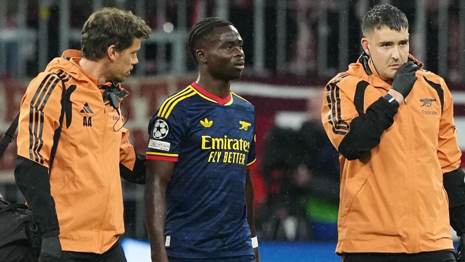 player in navy Arsenal kit walking off the pitch flanked by two staff members in orange jackets