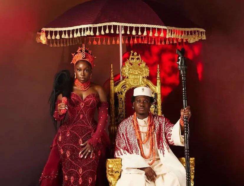 Bride in red beaded gown and groom in white agbada seated on a gold throne under a fringed umbrella
