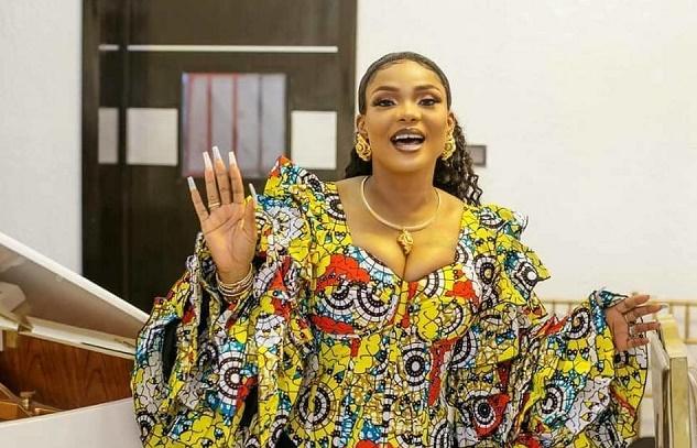 Woman in a colourful patterned dress with gold jewellery, smiling and waving, standing indoors beside a piano