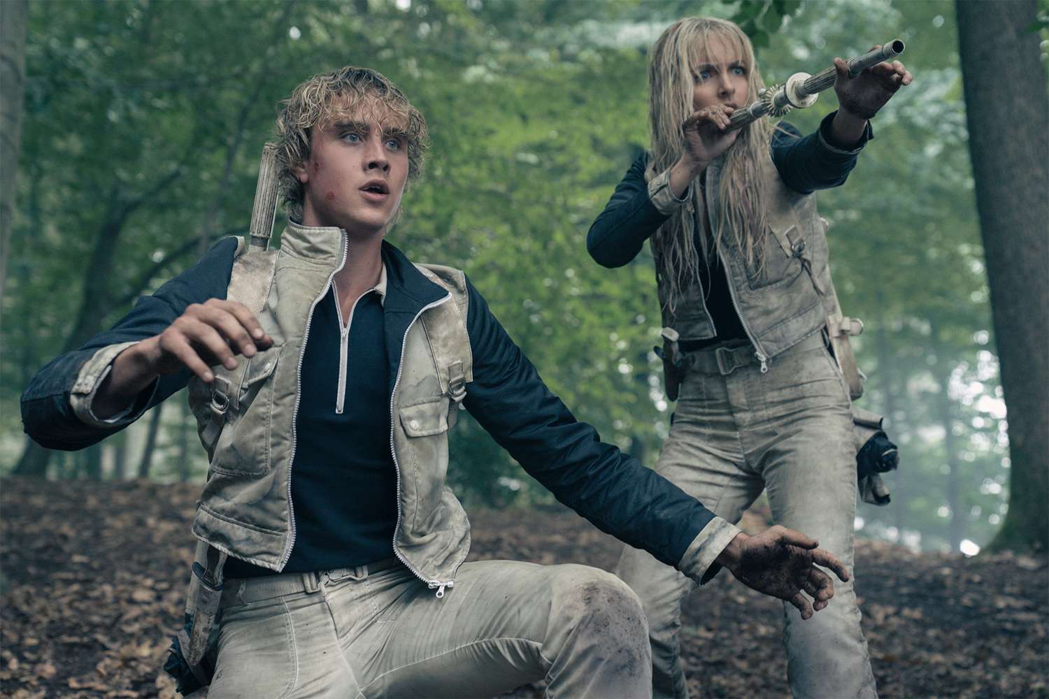 Young man crouching in a forest arena, another tribute aiming a slingshot behind him